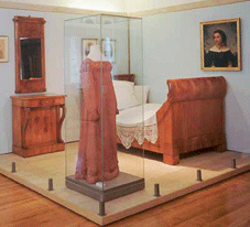 Die Abbildung zeigt einen Schlafraum und ei n Kleid in einer Glasvitrine aus der Ausstellung