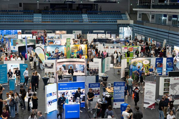 Eine große Messehalle mit zahlreichen Ständen und Bannern, die von verschiedenen Firmen gestaltet sind. Viele Menschen bewegen sich durch die Halle und interagieren an den Ständen.
