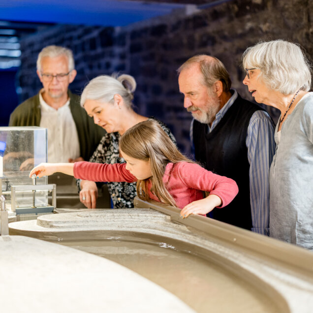 Eine Gruppe von fünf Personen, darunter ein Kind, betrachtet ein interaktives Modell mit Wasser in einer Ausstellung. Die Umgebung wirkt modern und beleuchtet.