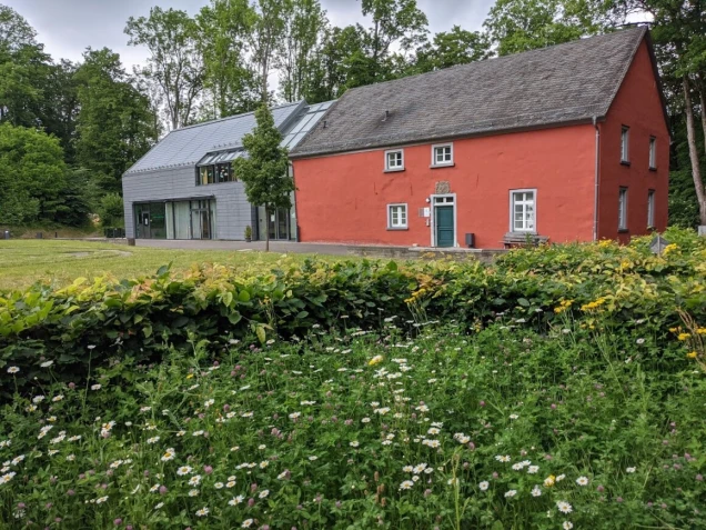 Ein rotes Gebäude mit grauem Dach steht neben einem modernen Anbau mit Glasfassade. Im Vordergrund sind eine Hecke und eine Wiese mit Wildblumen zu sehen. Dahinter befinden sich Bäume.
