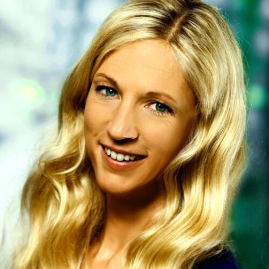Porträt einer lächelnden Person mit langen blonden Haaren, die wellig herabfallen. Die Person trägt ein dunkles Oberteil vor einem unscharfen Hintergrund.