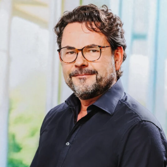 Ein Mann mit Brille, Bart und dunklem Hemd blickt lächelnd in die Kamera. Der Hintergrund ist unscharf und zeigt helle, moderne Strukturen.