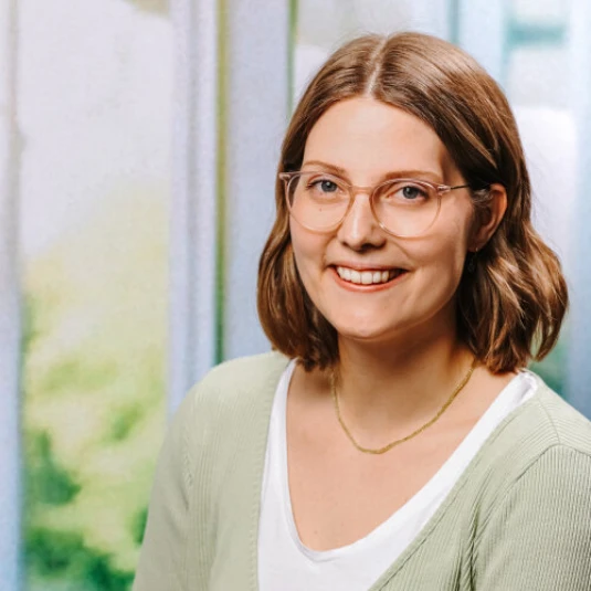 Eine lächelnde Person mit schulterlangem braunem Haar, Brille und grünem Oberteil vor einem unscharfen, hellen Hintergrund.