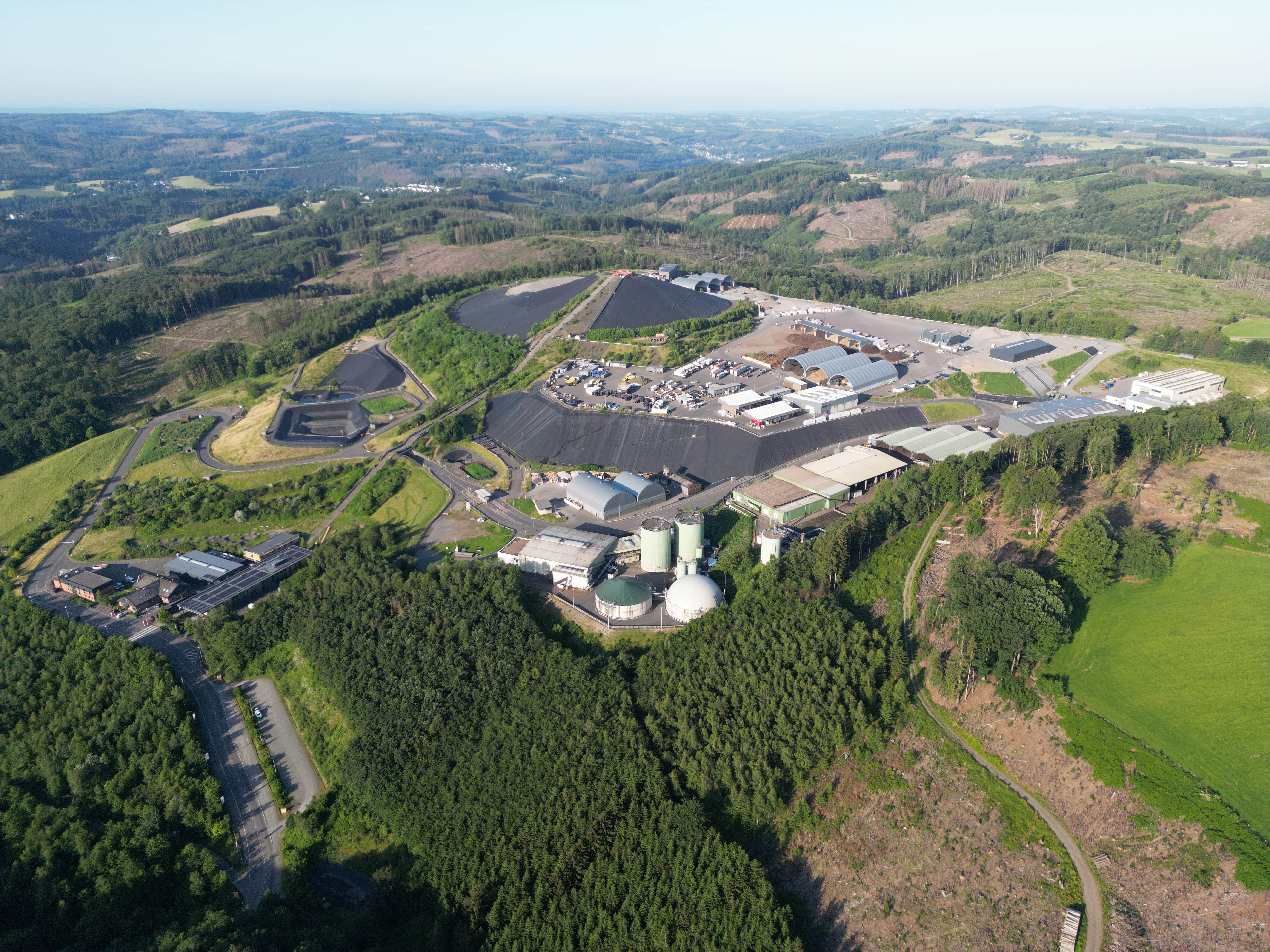 Luftaufnahme eines weitläufigen Industrie- und Recyclinggeländes mit Gebäuden, Silos und abgedeckten Bereichen, umgeben von grünen Wäldern und Feldern.