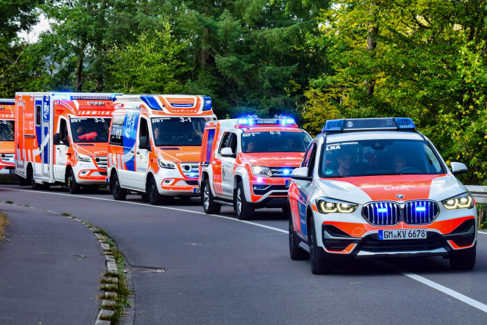 Eine Kolonne von Rettungsfahrzeugen, darunter ein BMW-SUV und mehrere Krankenwagen, fährt mit eingeschaltetem Blaulicht auf einer Straße durch eine grüne Umgebung.