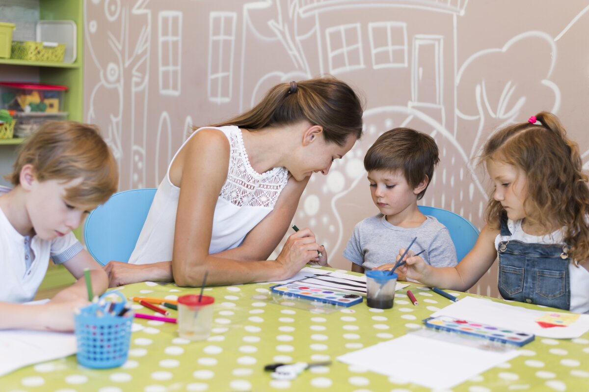 Eine Frau sitzt mit drei Kindern an einem Tisch, der mit einer grünen Tischdecke mit weißen Punkten bedeckt ist. Sie malen mit Wasserfarben und Stiften. Im Hintergrund ist eine Wand mit gemalten weißen Haus- und Baumumrissen zu sehen.