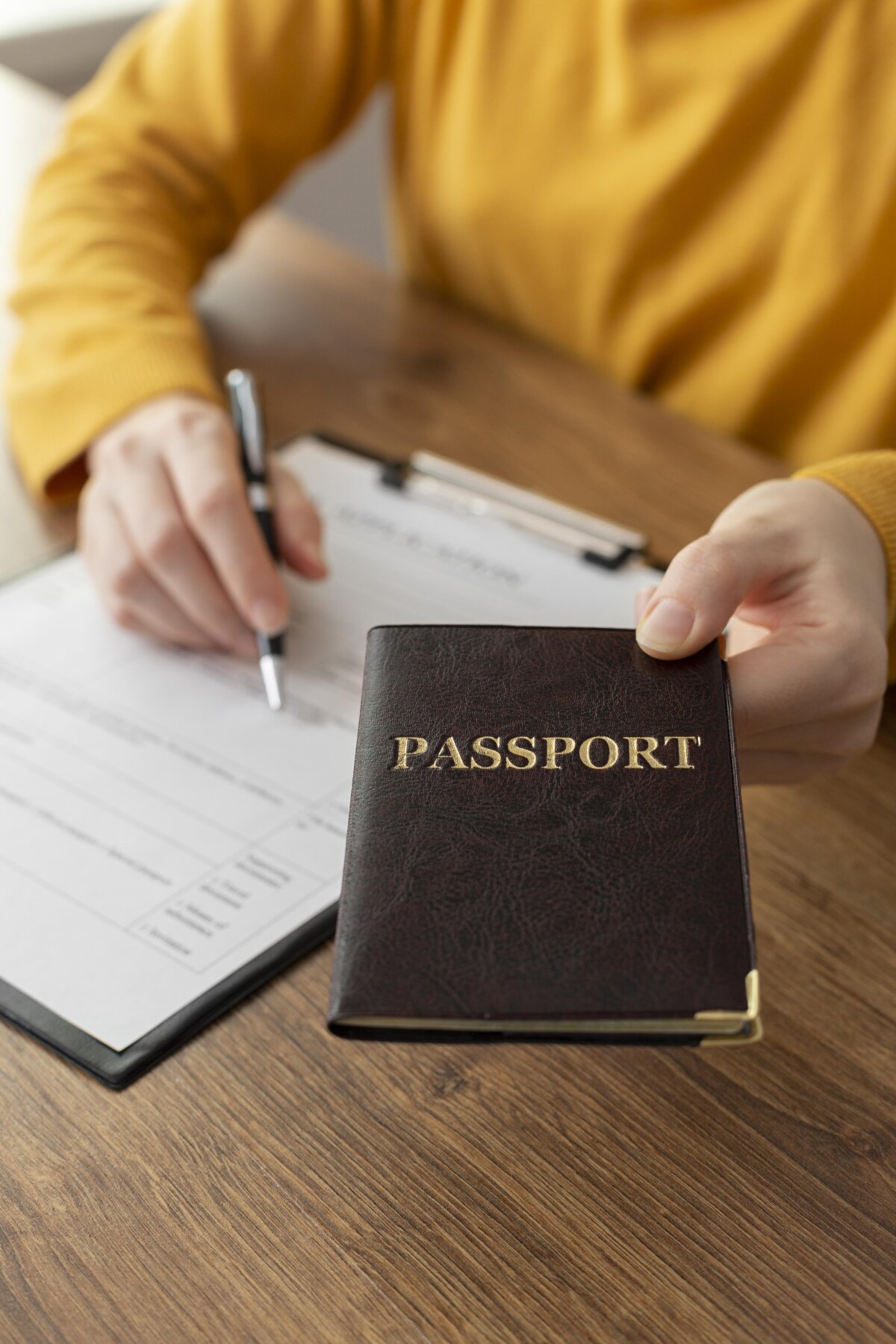 Eine Person in gelber Kleidung hält einen dunklen Reisepass mit goldener Aufschrift 'PASSPORT' in der Hand. Im Hintergrund füllt sie ein Formular auf einem Klemmbrett aus.