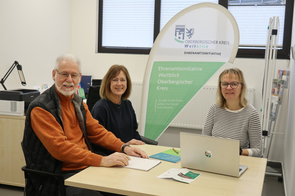 Drei Personen sitzen an einem Tisch in einem Büro. Ein Mann und zwei Frauen lächeln in die Kamera. Im Hintergrund ein Banner der Ehrenamtsinitiative Weitblick Oberbergischer Kreis sowie ein Fenster.