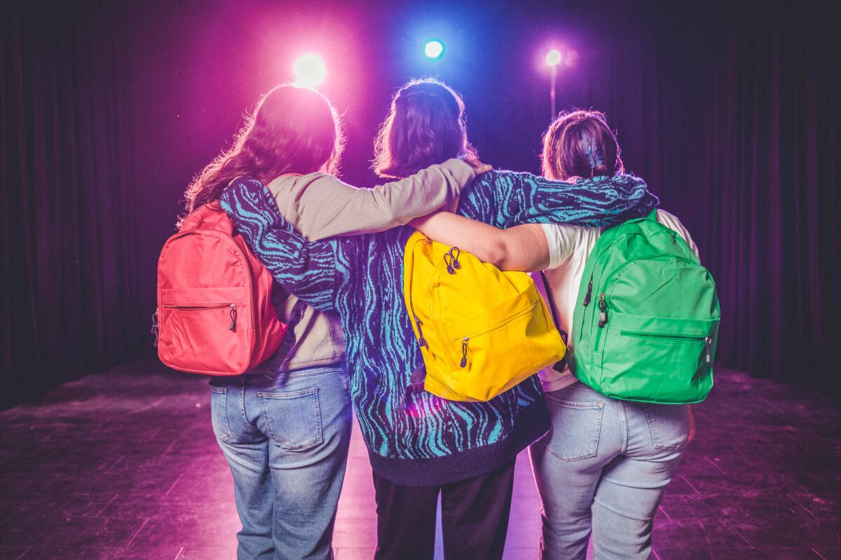 Drei Personen stehen Rücken zur Kamera, Arme umeinander gelegt. Jede trägt einen bunten Rucksack (rot, gelb, grün). Im Hintergrund bunte Lichter und ein schwarzer Vorhang.