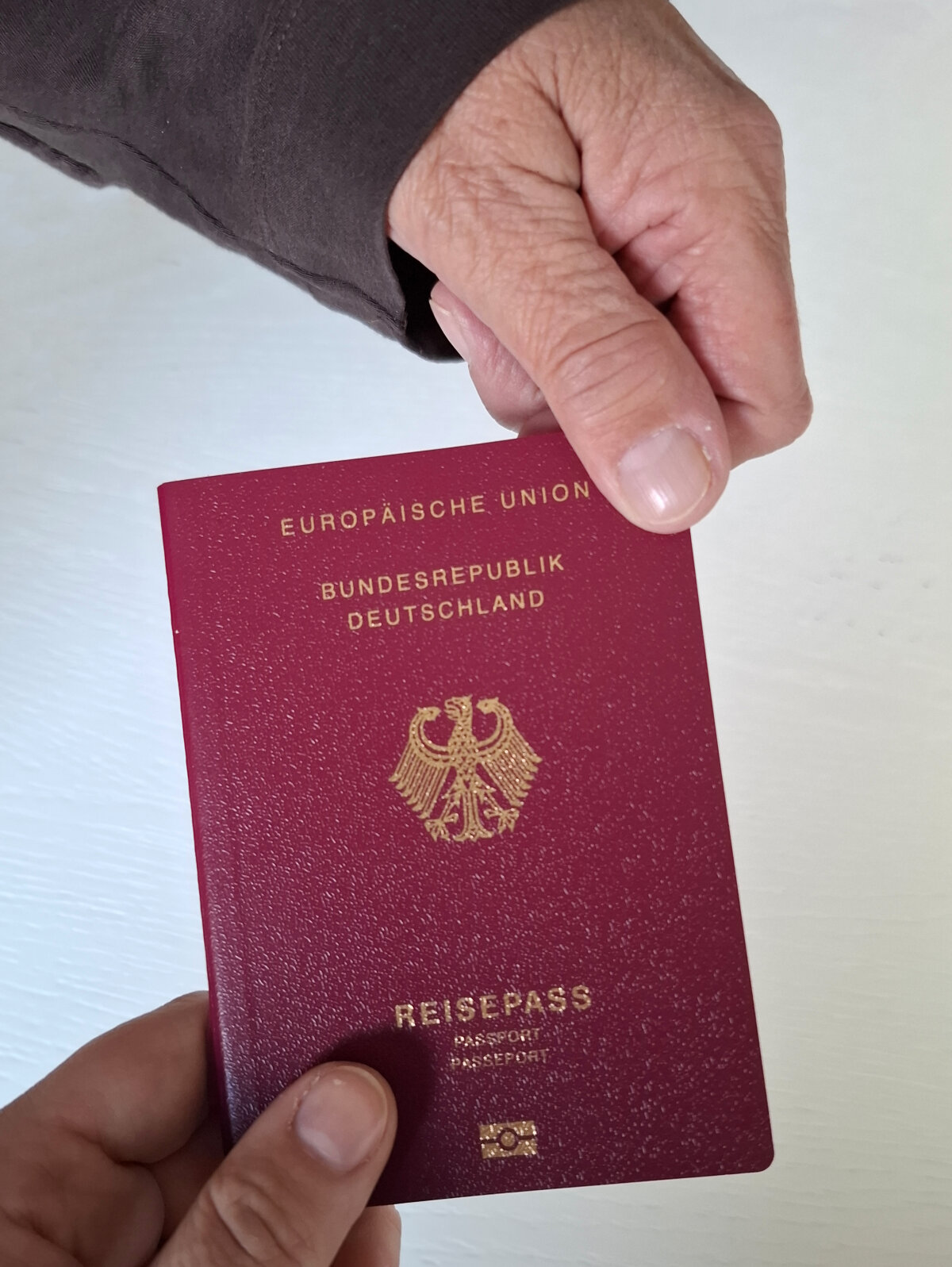 Zwei Hände halten gemeinsam einen deutschen Reisepass mit bordeauxrotem Einband und goldener Prägung, auf der der Bundesadler zu sehen ist.