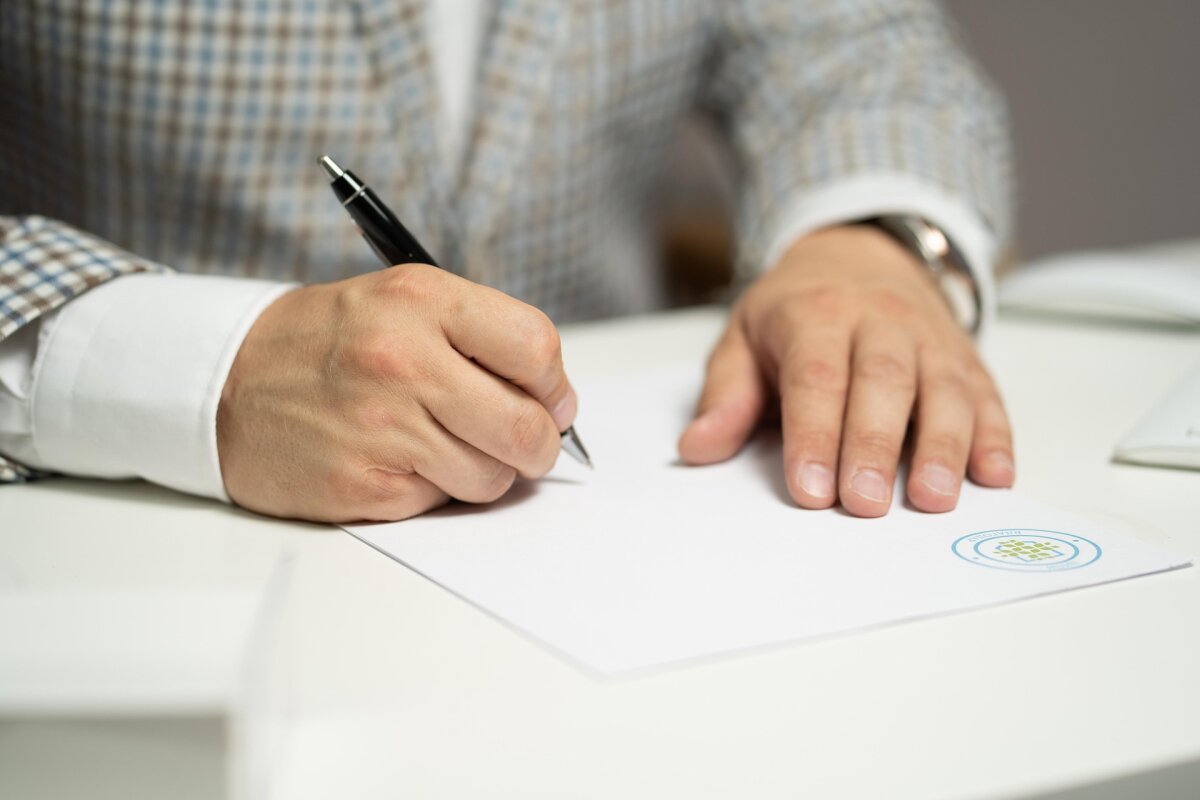 Eine Person in einem karierten Sakko schreibt mit einem schwarzen Stift auf ein weißes Blatt Papier, das auf einem Tisch liegt. Auf dem Papier ist ein blau-grünes Logo erkennbar.