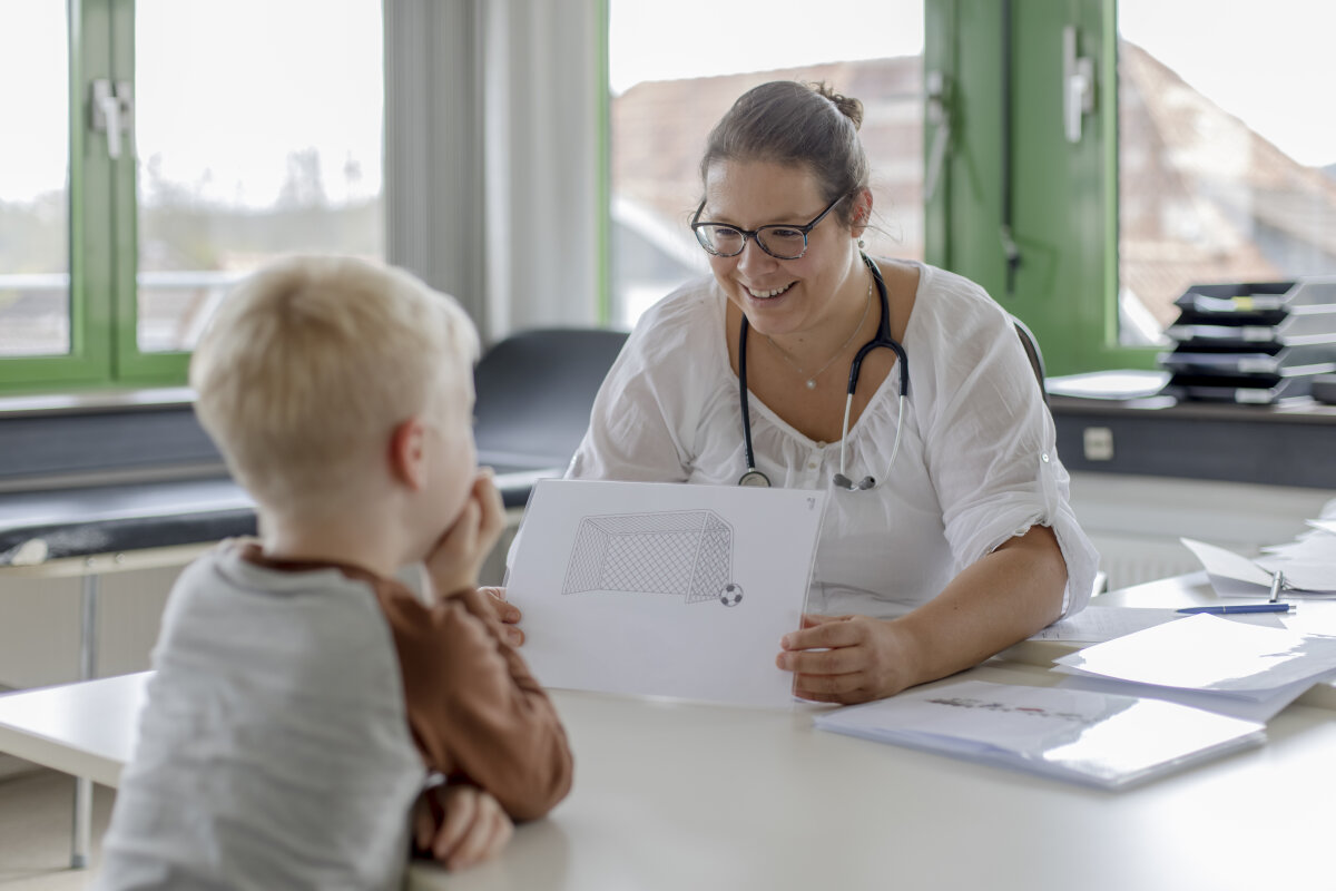 Eine Frau mit Brille und Stethoskop sitzt an einem Schreibtisch und zeigt einem Kind mit blondem Haar eine Zeichnung von einem Fußballtor. Im Hintergrund sind grüne Fensterrahmen zu sehen.
