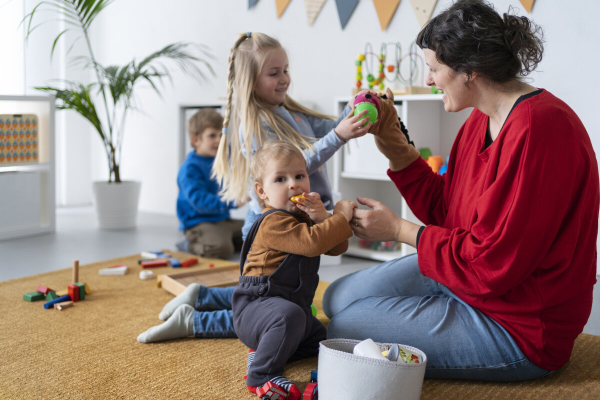 Eine Frau in rotem Oberteil spielt mit drei Kindern in einem hellen Raum. Spielzeug ist auf einem Teppich verteilt, und im Hintergrund stehen Regale mit weiteren Spielsachen.