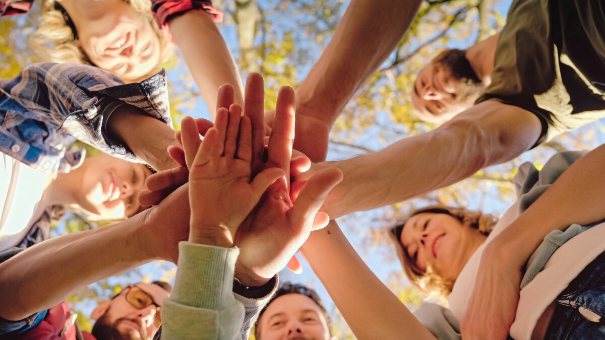 Gruppe von Menschen, die im Kreis stehen und ihre Hände in der Mitte übereinanderlegen, mit Bäumen und blauem Himmel im Hintergrund.