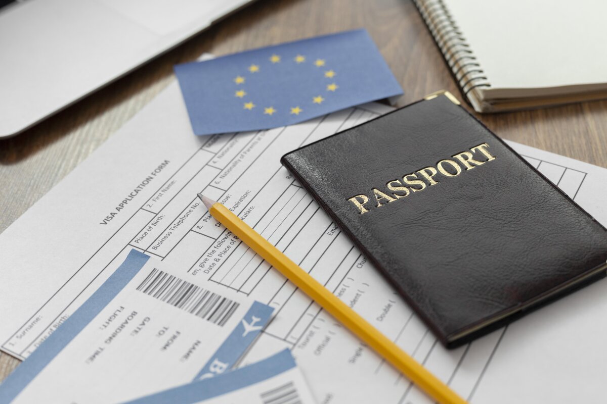 Ein brauner Reisepass, ein gelber Bleistift, ein Visa-Antragsformular, Flugtickets und eine EU-Flagge liegen auf einem Holztisch. Im Hintergrund ein Laptop und ein Notizbuch.