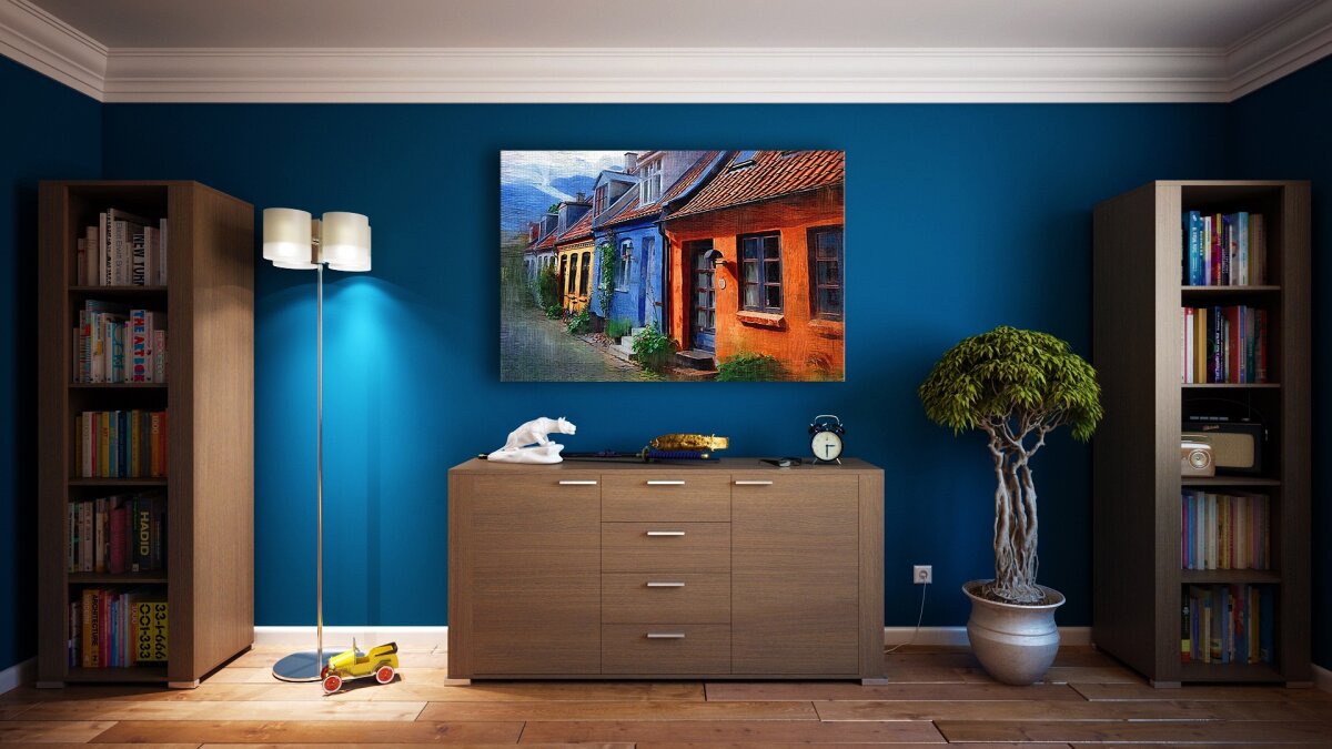 Ein Wohnzimmer mit blauer Wand, zwei Bücherregalen, einer Holzkommode, einer Stehlampe, einem gerahmten Gemälde, einer Zimmerpflanze und Dekoobjekten.