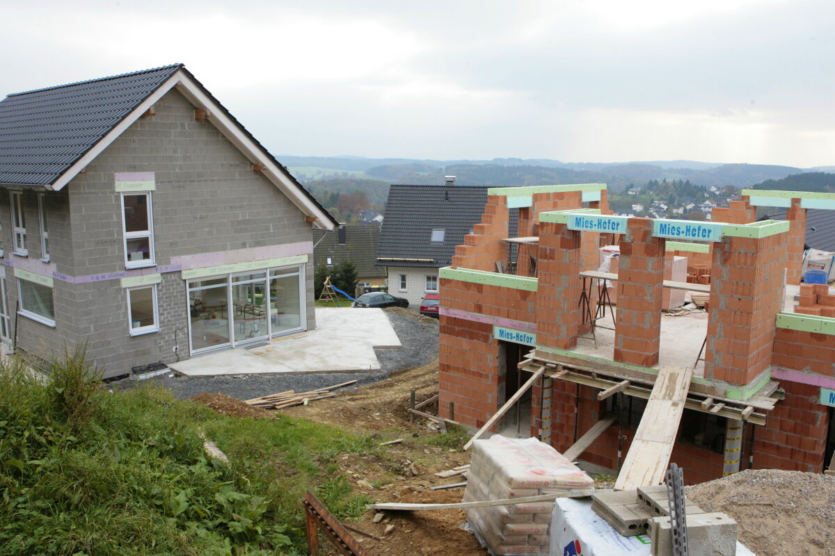 Eine Baustelle mit einem teilweise fertiggestellten Haus aus Ziegeln rechts und einem fast fertigen Haus mit grauer Fassade links. Im Hintergrund eine hügelige Landschaft.