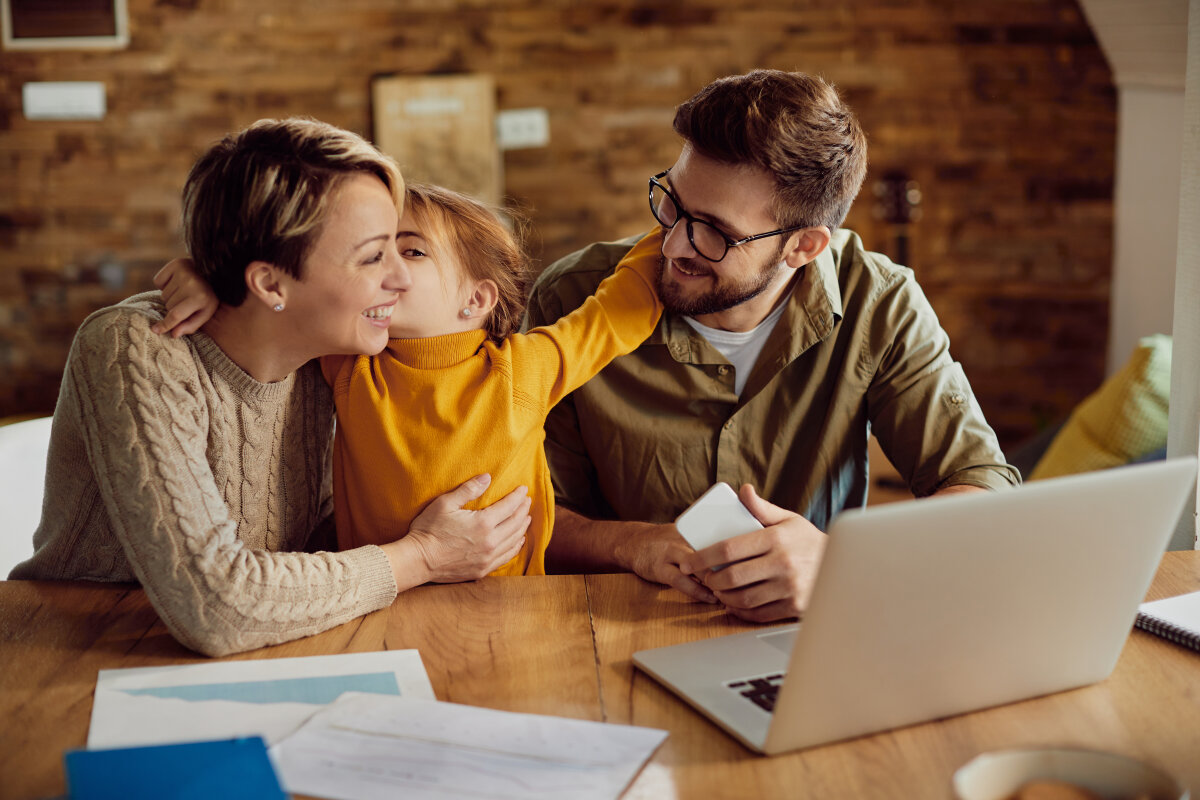 Ein fröhliches Familienbild: Eine Frau, ein Mann und ein Kind sitzen an einem Tisch. Die Frau und das Kind umarmen sich, während der Mann lächelnd zusieht. Auf dem Tisch liegen Dokumente und ein Laptop.