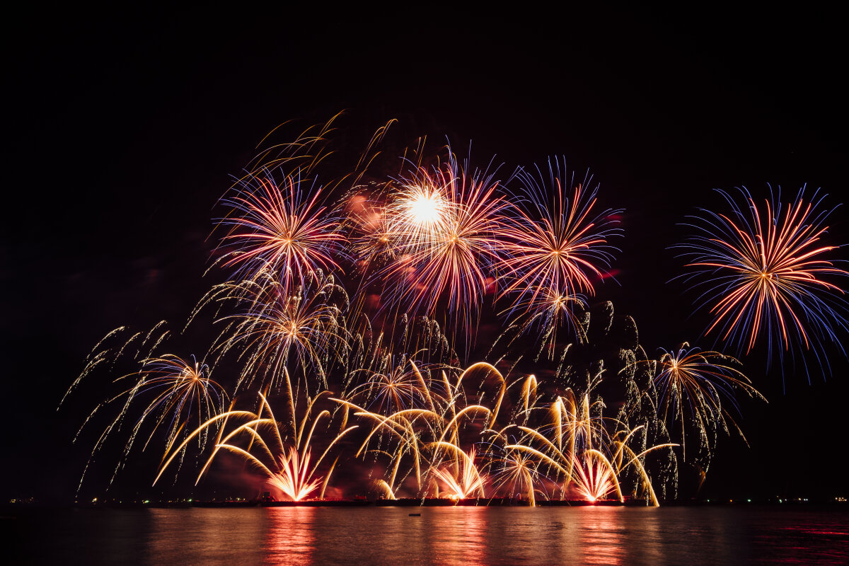 Ein nächtlicher Himmel mit buntem Feuerwerk in Rot, Gelb und Blau, das über einem Gewässer explodiert. Die Reflexionen sind im Wasser sichtbar.