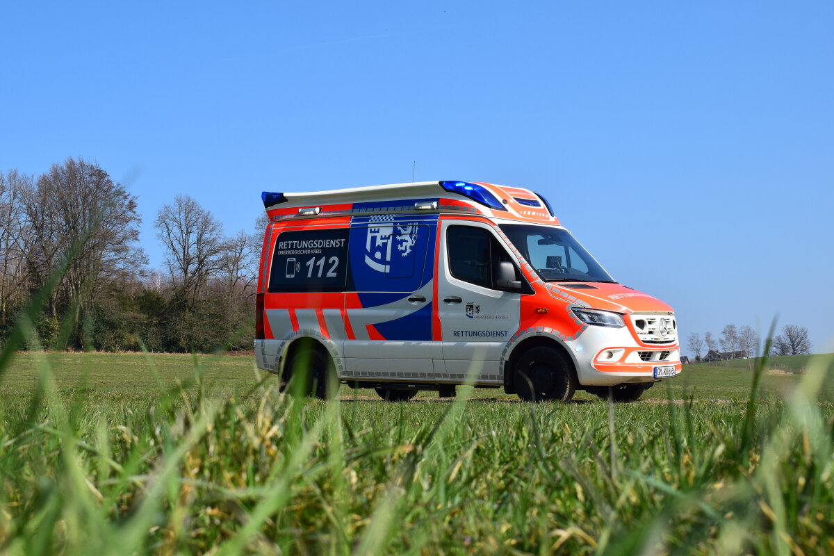 Ein Rettungswagen mit orange-weißer Lackierung und blauen Akzenten steht auf einer Wiese. Im Hintergrund sind Bäume und ein klarer blauer Himmel sichtbar.