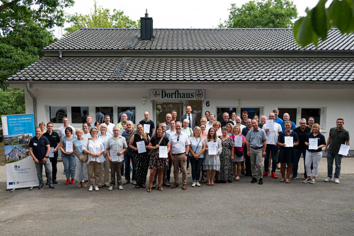 Eine Gruppe von Menschen posiert vor einem Gebäude mit der Aufschrift 'Dorhaus'. Einige Personen halten Urkunden in den Händen. Im Hintergrund sind Bäume sichtbar.