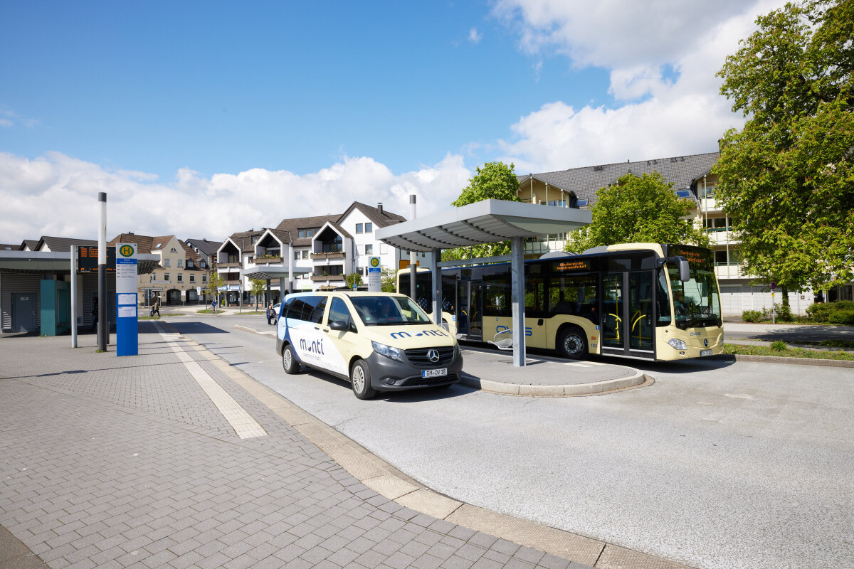 Eine Bushaltestelle mit einem beige-gelben Linienbus und einem beige-blauen Kleinbus mit der Aufschrift 'monti', umgeben von modernen Wohnhäusern, Bäumen und klarem Himmel.