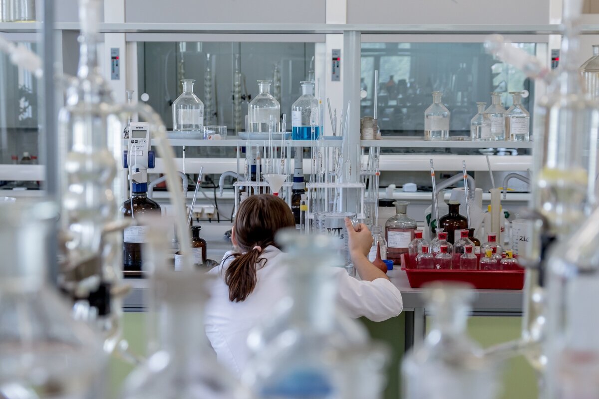 Eine Person in einem weißen Laborkittel arbeitet in einem Labor, umgeben von chemischen Glasgefäßen, Reagenzflaschen und Pipetten auf einem Labortisch.