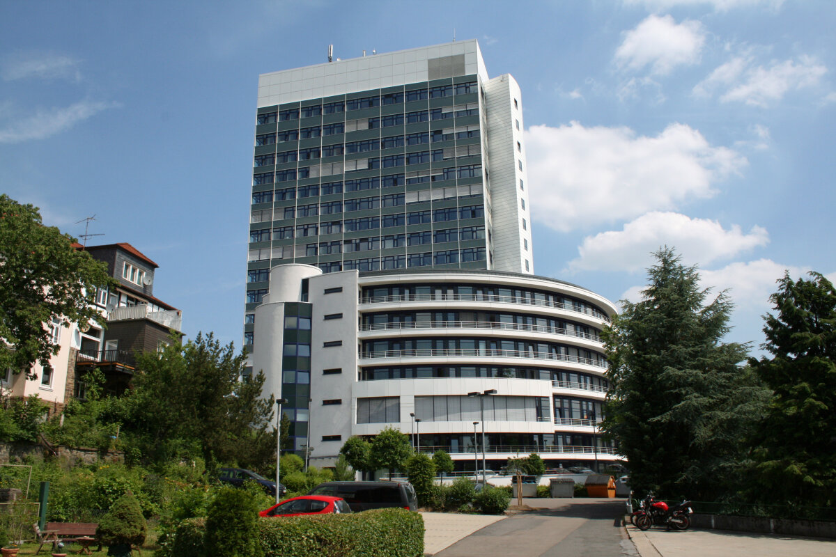 Mehrstöckiges modernes Gebäude mit Glasfassade und rundem Anbau, umgeben von Bäumen, einem Gehweg und geparkten Fahrzeugen unter blauem Himmel.
