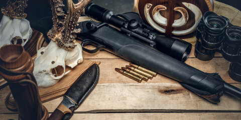 Auf einem Holztisch liegen ein Gewehr mit Zielfernrohr, Patronen, ein Jagdmesser in einer Lederscheide, ein Fernglas, Hirschgeweihe und Tierknochen.