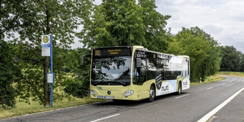 Ein beiger Linienbus mit der Nummer 303 und Zielort Gummersbach Bahnhof steht an einer ländlichen Bushaltestelle. Im Hintergrund sind Bäume und eine Straße sichtbar.