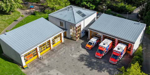 Luftaufnahme einer Rettungswache mit zwei Gebäuden. Vor einer Garage stehen drei Einsatzfahrzeuge. Umgeben von Grünflächen und Bäumen.