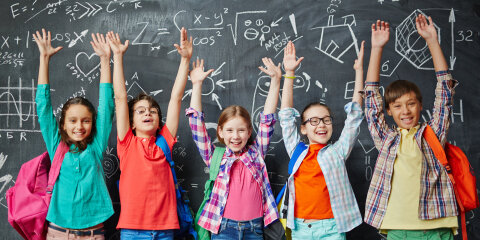 Fünf Kinder mit bunten Kleidern und Rucksäcken stehen vor einer Tafel mit mathematischen Formeln und heben fröhlich die Arme.