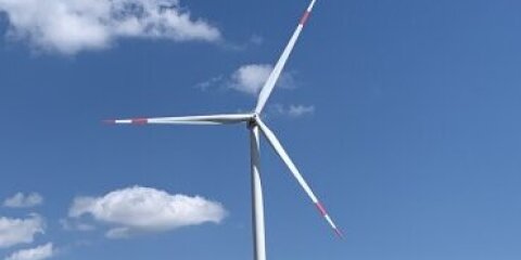 Mehrere Windkraftanlagen stehen in einer offenen Landschaft mit trockenen Wiesen und vereinzelten Bäumen im Hintergrund unter einem blauen Himmel mit wenigen Wolken.