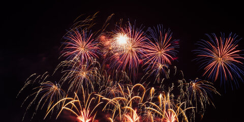 Ein nächtlicher Himmel mit buntem Feuerwerk in Rot, Gelb und Blau, das über einem Gewässer explodiert. Die Reflexionen sind im Wasser sichtbar.