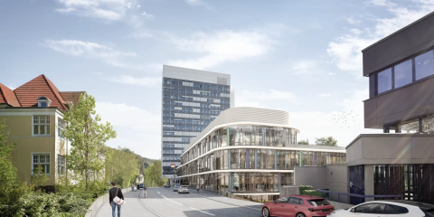 Städtische Straßenszene mit modernen und historischen Gebäuden. Im Vordergrund parken Autos, eine Person geht auf dem Gehweg, und im Hintergrund ist ein hoher Bürokomplex zu sehen.