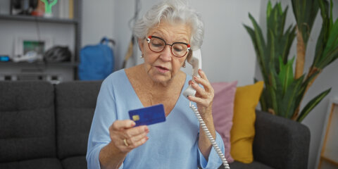 Eine ältere Frau mit Brille und grauen Haaren sitzt auf einem Sofa und telefoniert mit einem Festnetztelefon. In ihrer Hand hält sie eine blaue Kreditkarte. Im Hintergrund sind ein Regal, Kissen und eine Zimmerpflanze zu sehen.