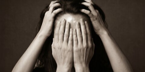Eine Person hält sich mit zwei Händen den Kopf, während zwei andere Hände das Gesicht bedecken. Der Hintergrund ist dunkel und einfarbig.