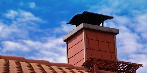 Rotes Ziegeldach mit einem roten Kamin, der eine schwarze Abdeckung hat. Der Himmel ist blau mit weißen Wolken.