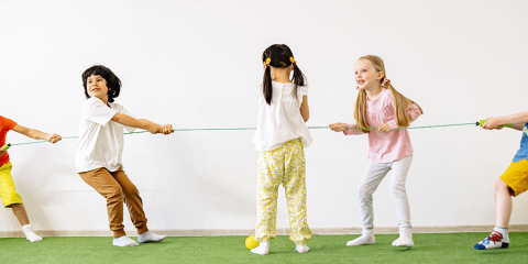 Kinder spielen Tauziehen auf grünem Boden in einem hellen Raum. Zwei Teams ziehen an einem Seil, jedes Team besteht aus zwei Kindern, ein Kind steht in der Mitte.