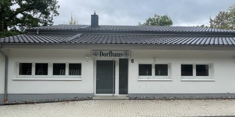 Ein einstöckiges Gebäude mit weißer Fassade und einem schwarzen Ziegeldach. Über der Eingangstür steht 'Dorfhaus'. Links und rechts befinden sich Fenster.