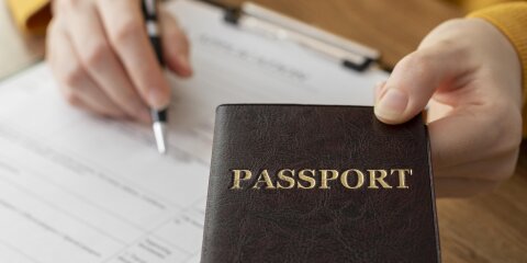 Eine Person in gelber Kleidung hält einen dunklen Reisepass mit goldener Aufschrift 'PASSPORT' in der Hand. Im Hintergrund füllt sie ein Formular auf einem Klemmbrett aus.
