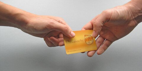 Zwei Hände halten gemeinsam eine orangefarbene Karte mit der Aufschrift 'Ehrensache' und einem Wappen. Der Hintergrund ist grau.