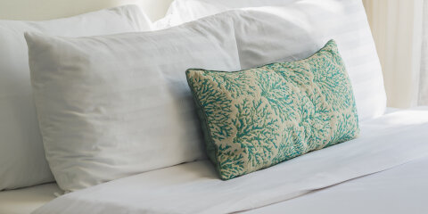 Ein Bett mit weißer Bettwäsche, zwei große weiße Kissen und einem kleineren Kissen mit grünem Blattmuster. Eine Wandleuchte ist daneben sichtbar.