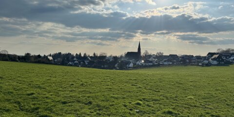 Grüne Wiese im Vordergrund, dahinter eine Siedlung mit Häusern und einer Kirche mit spitzem Turm. Der Himmel ist teils bewölkt mit Sonnenstrahlen.
