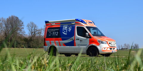 Ein Rettungswagen mit orange-weißer Lackierung und blauen Akzenten steht auf einer Wiese. Im Hintergrund sind Bäume und ein klarer blauer Himmel sichtbar.