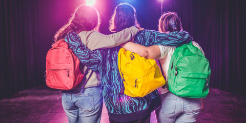 Drei Personen stehen Rücken zur Kamera, Arme umeinander gelegt. Jede trägt einen bunten Rucksack (rot, gelb, grün). Im Hintergrund bunte Lichter und ein schwarzer Vorhang.