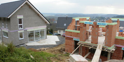 Eine Baustelle mit einem fast fertigen Einfamilienhaus aus grauen Steinen und einem im Bau befindlichen Gebäude aus roten Ziegelsteinen. Im Hintergrund sind weitere Häuser und eine hügelige Landschaft zu sehen.