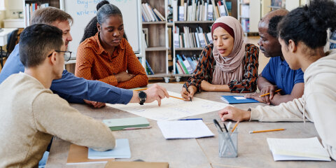 Eine Gruppe von sechs Menschen sitzt an einem Tisch in einer Bibliothek und arbeitet zusammen an einem großen Blatt Papier mit Notizen. Im Hintergrund stehen Bücherregale und eine Tafel mit englischen Verben.