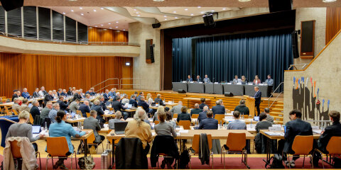 Ein großer Konferenzraum mit Holzverkleidung, gefüllt mit Menschen an Tischen, die einer Podiumsdiskussion vor einer Bühne mit mehreren Rednern folgen.