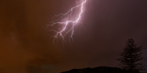 Ein Blitz erhellt den Nachthimmel über einer dunklen Landschaft mit Bäumen und Hügeln. Ein einzelner Baum ist rechts im Vordergrund zu sehen.